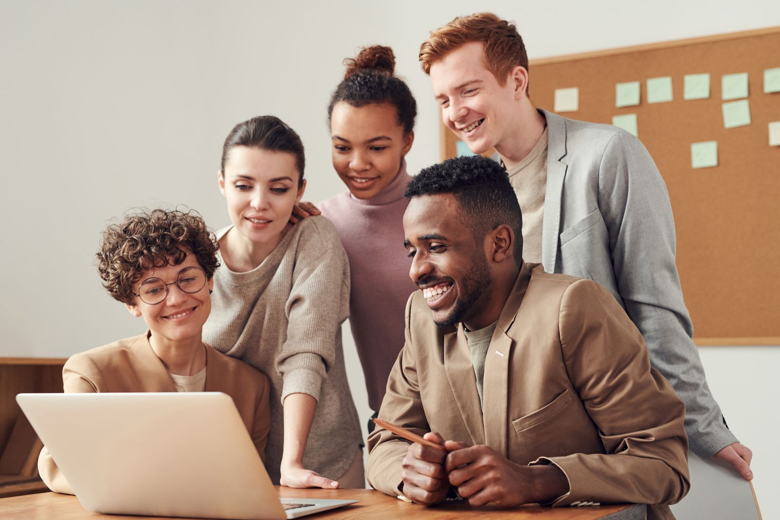 Cinq personnes se réunissent autour d'un ordinateur portable, souriant en explorant comment trouver un stage rapidement dans un espace de bureau lumineux avec un tableau en liège et des notes autocollantes en arrière-plan.