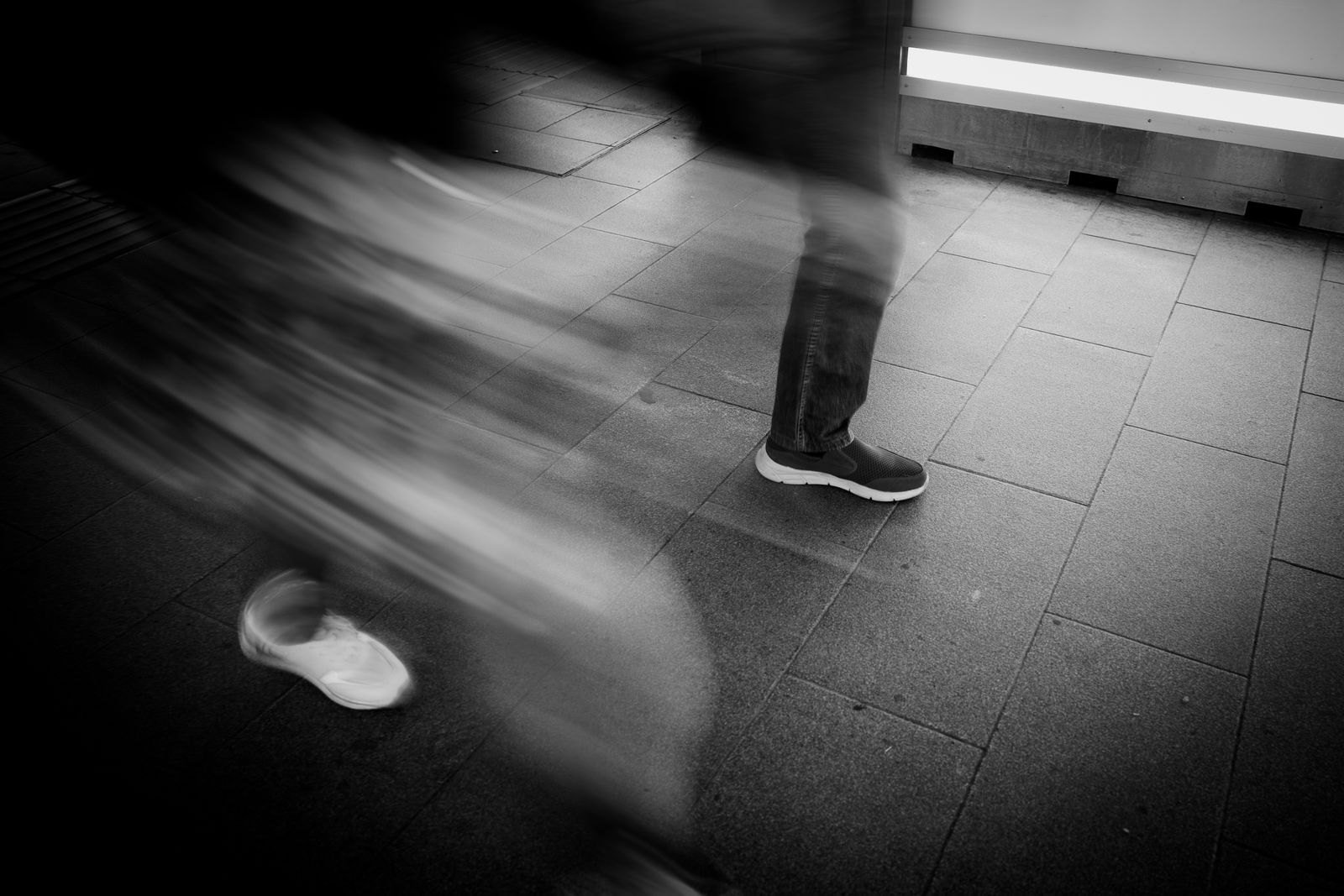 Photo en noir et blanc de personnages flous en mouvement marchant sur un trottoir carrelé ; seuls le bas des jambes et les pieds sont visibles, ce qui met l'accent sur le mouvement et la vitesse - en écho à la précipitation de l'alternance et de l'urgence.