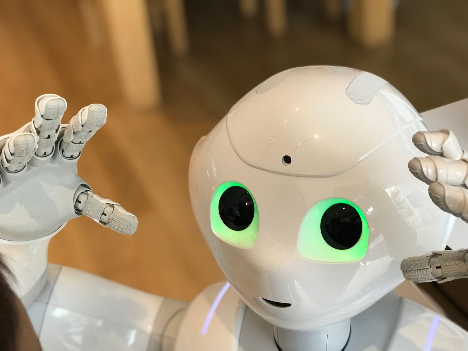 Un robot humanoïde symbolisant un ATS examinant un CV et son format aux yeux verts luisants lève les mains, paumes vers l'avant. Avec son extérieur blanc et lisse et son visage amical et expressif, il se tient sur un sol en bois, comme un CV ATS conçu pour une interaction transparente.