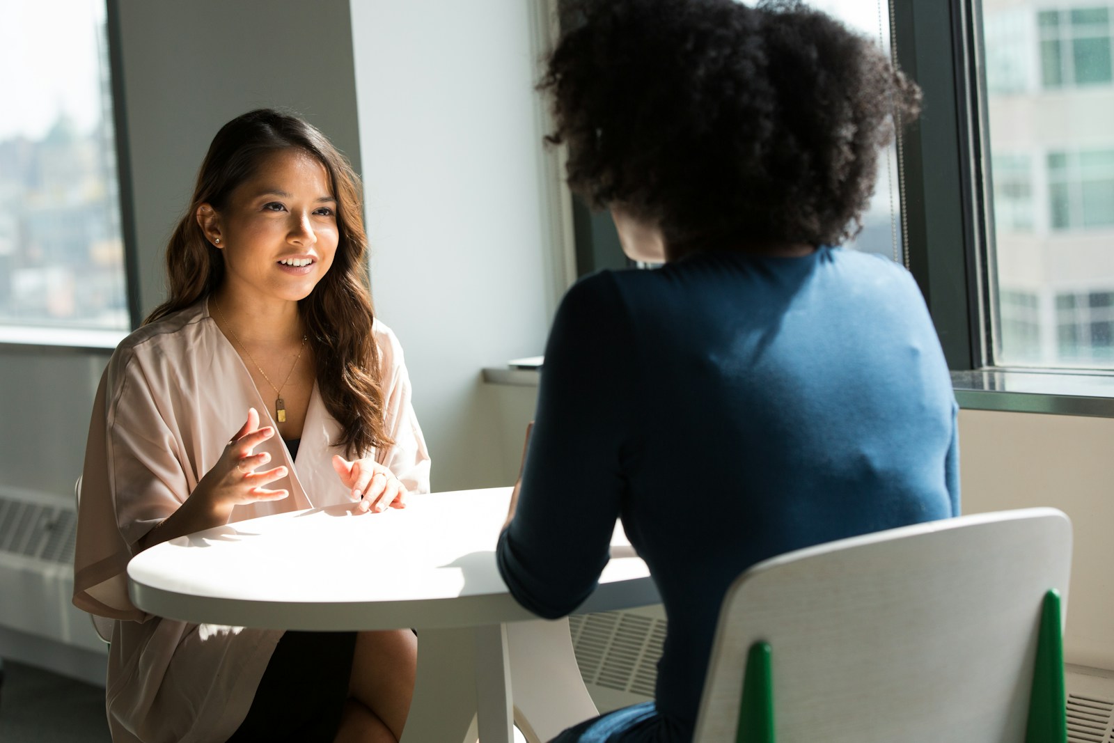Deux femmes sont assises face à face à une table ronde près d'une fenêtre, apparemment en train de mener un entretien de stage étudiant : l'une parle et gesticule, tandis que l'autre l'écoute attentivement, dos à la caméra.